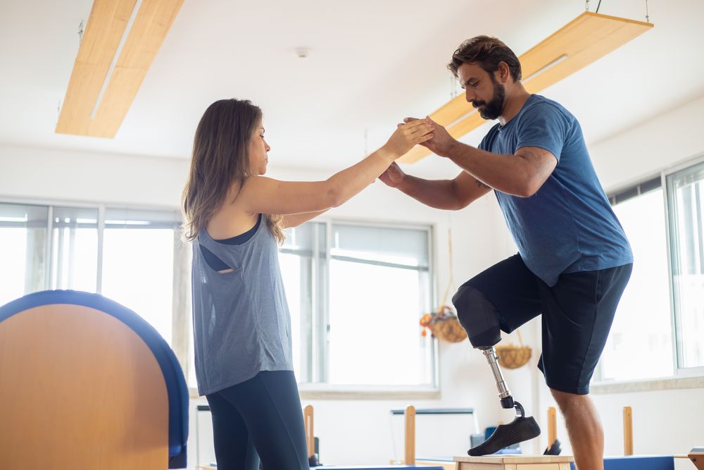 Fisioterapeuta sostiene las manos de un paciente con prótesis de pierna durante un ejercicio de equilibrio en el gimnasio del centro.